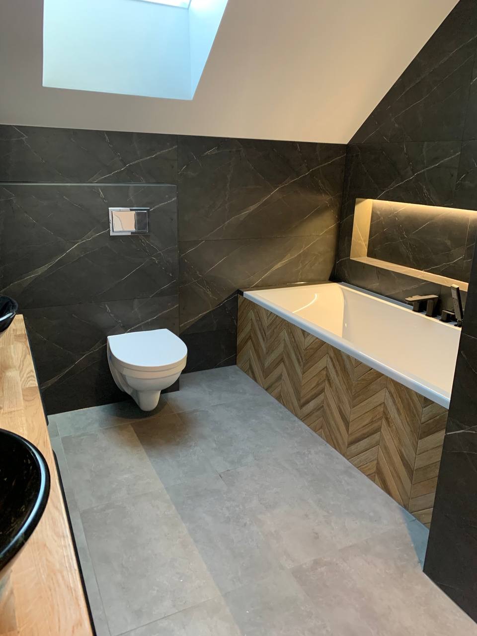 Nowoczesna łazienka z czarnymi płytkami imitującymi marmur, podwieszaną toaletą, wanną obudowaną drewnianą mozaiką w jodełkę, szarą podłogą i oknem dachowym w skosie sufitu.
