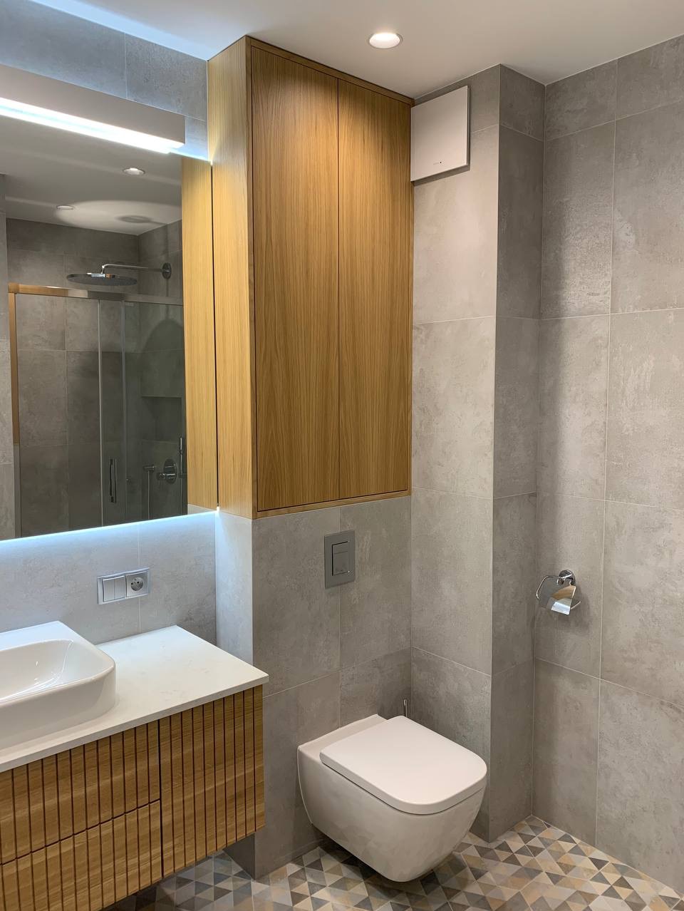 Nowoczesna łazienka z szarą płytką, podwieszaną toaletą, umywalką nablatową na szafce z drewnianymi lamelami i szafką wiszącą z drewna, z geometryczną mozaiką na podłodze.