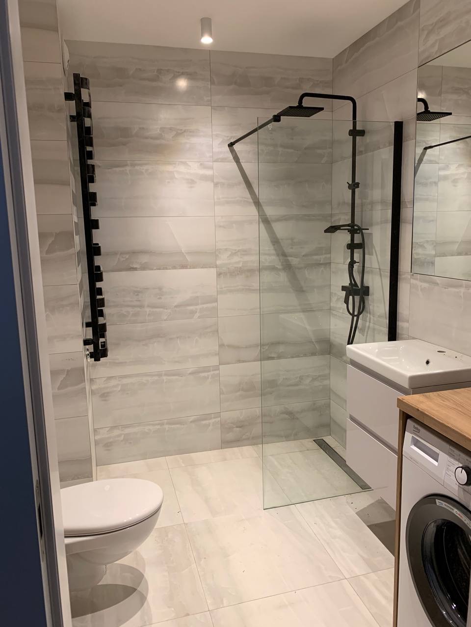 Nowoczesna łazienka z kabiną prysznicową typu walk-in z czarną armaturą, grzejnikiem drabinkowym i pralką pod blatem umywalki, wykończona płytkami imitującymi marmur.