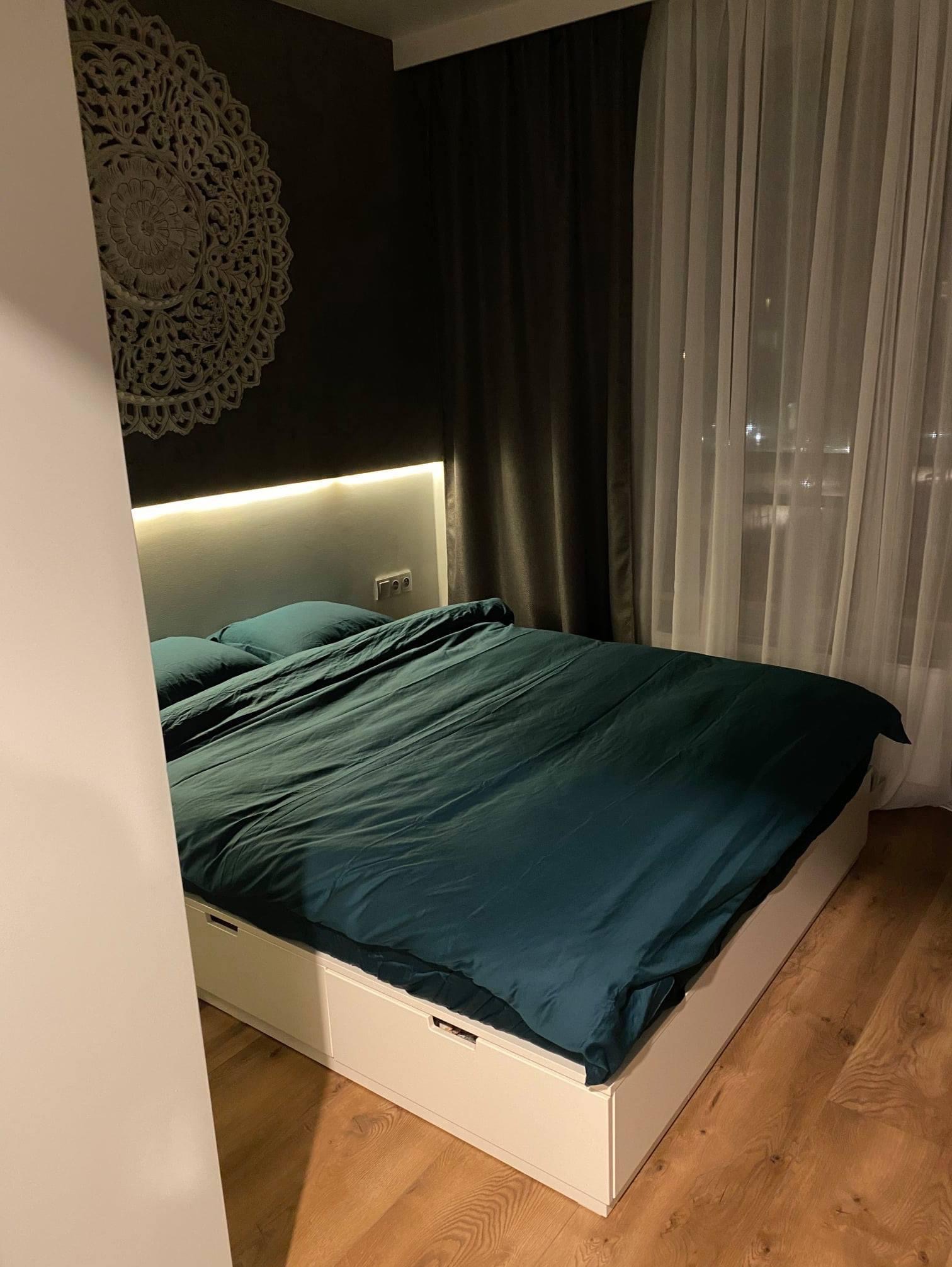 Sypialnia z łóżkiem z szufladami, wezgłowiem podświetlonym LED, dekoracyjną rozetą na ciemnej ścianie i zasłonami w oknie.