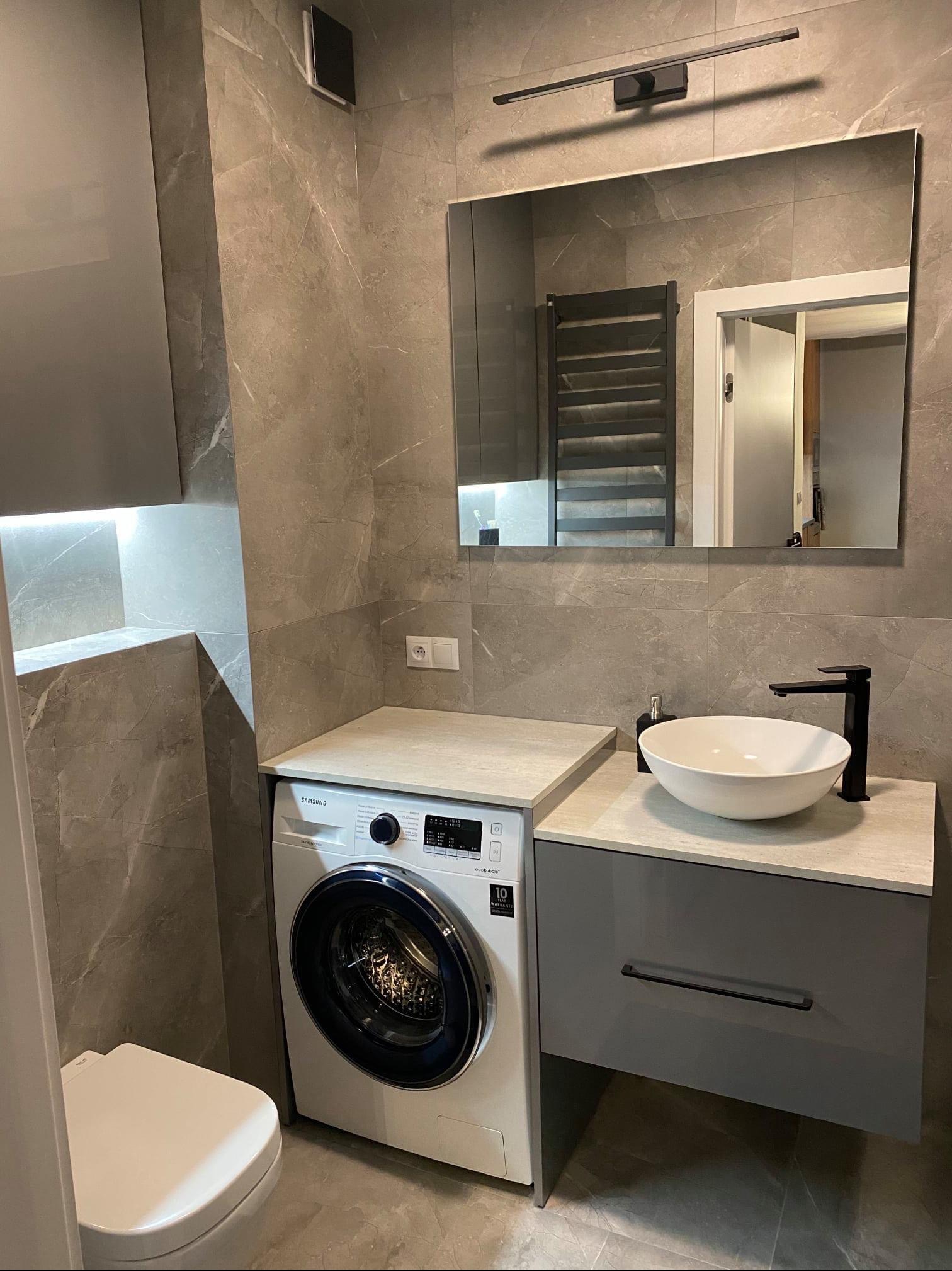Nowoczesna łazienka w szarościach z marmurkowymi płytkami, umywalką nablatową, zabudową pralki i minimalistycznym oświetleniem LED nad lustrem.