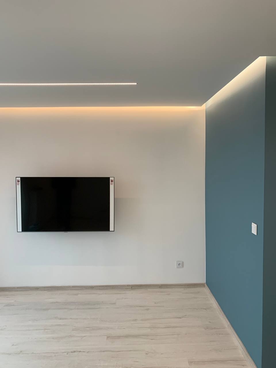 Minimalistyczne wnętrze po remoncie: białe ściany, jedna ściana pomalowana na niebiesko, podwieszony telewizor, panele podłogowe w odcieniach szarości i białe listwy przypodłogowe, oświetlenie LED...