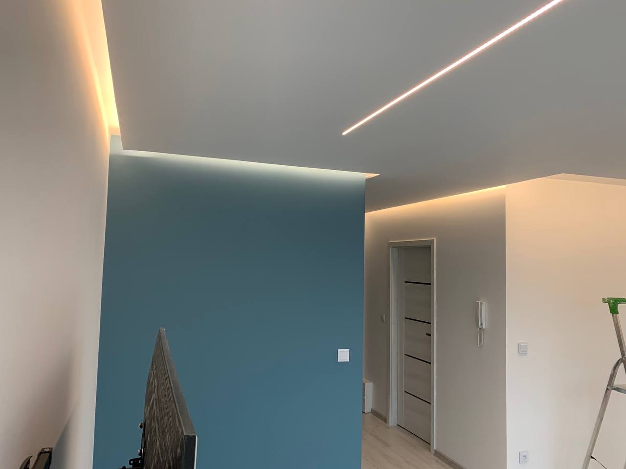 Nowoczesne wnętrze po odświeżeniu: błękitna ściana kontrastuje z białymi ścianami i sufitem z liniowym oświetleniem LED; widoczna drabina malarska w rogu.