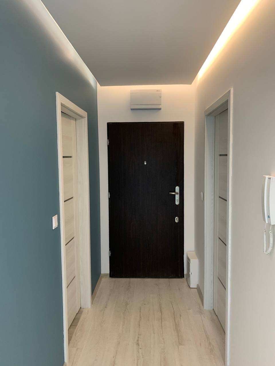 Korytarz po odświeżeniu z ciemnymi drzwiami wejściowymi, panelami podłogowymi w odcieniach beżu, pomalowaną na niebiesko ścianą po lewej i oświetleniem LED w suficie podwieszanym.