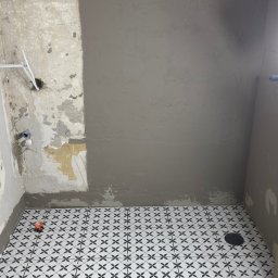 DINAS MASTER DMYTRO OKHRIMENKO - Łazienka w trakcie prac: ściana częściowo pokryta szarą gładzią, podłoga wyłożona biało-czarną mozaiką, widoczne narzędzia glazurnicze i fragment instalacji wodnej.