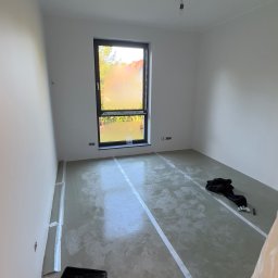 DINAS MASTER DMYTRO OKHRIMENKO - Przygotowane podłoże pod wylewkę samopoziomującą, zabezpieczone taśmą przy ścianach, widoczna kuweta malarska i ubrania robocze w pustym pomieszczeniu z oknem.