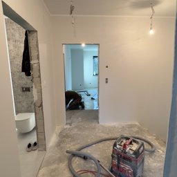 DINAS MASTER DMYTRO OKHRIMENKO - Wnętrze w trakcie prac wykończeniowych: widoczny odkurzacz przemysłowy, surowy beton na podłodze, ściany po nałożeniu gładzi, oraz fragment łazienki z toaletą w tle. W głębi pomieszczenia, osoba...
