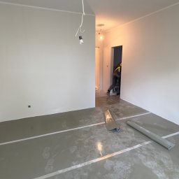 DINAS MASTER DMYTRO OKHRIMENKO - Przygotowanie do prac wykończeniowych w pustym pomieszczeniu, podłoga pokryta wylewką z widocznymi śladami butów, zabezpieczona folią malarską i taśmami, zwisający kabel z żarówką. W tle widoczna...