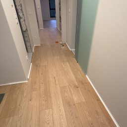 DINAS MASTER DMYTRO OKHRIMENKO - Nowo ułożona, jasna podłoga panelowa w korytarzu z widocznym fragmentem ściany w stanie surowym i zamontowanymi przesuwnymi drzwiami.