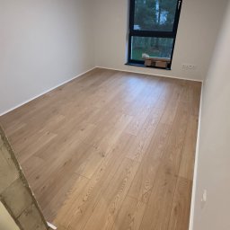 DINAS MASTER DMYTRO OKHRIMENKO - Jasne panele podłogowe ułożone w nowym pomieszczeniu z oknem, widoczne kartony pod oknem, surowy betonowy element konstrukcyjny w lewym dolnym rogu.