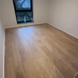 Jasne panele podłogowe ułożone w pokoju z oknem w kształcie trapezu, widok na dom z ogrodem, białe ściany, listwy przypodłogowe, butelka wody i silikon przy oknie.