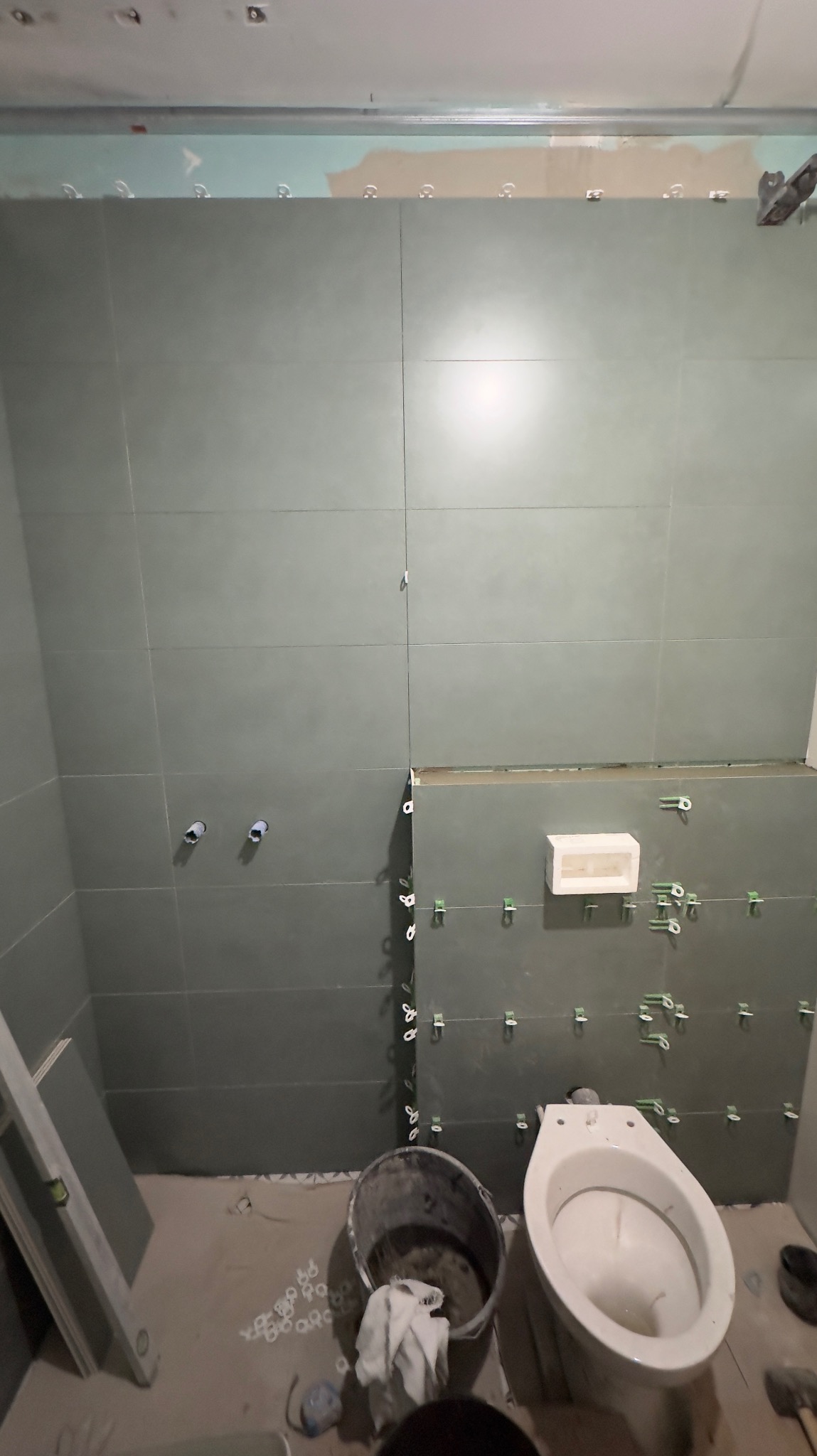 Świeżo wyłożone, szare płytki na ścianie łazienki z widocznymi krzyżykami dystansowymi, biała toaleta i wiadro z zaprawą, poziomica oparta o ścianę, przygotowane do dalszych prac.