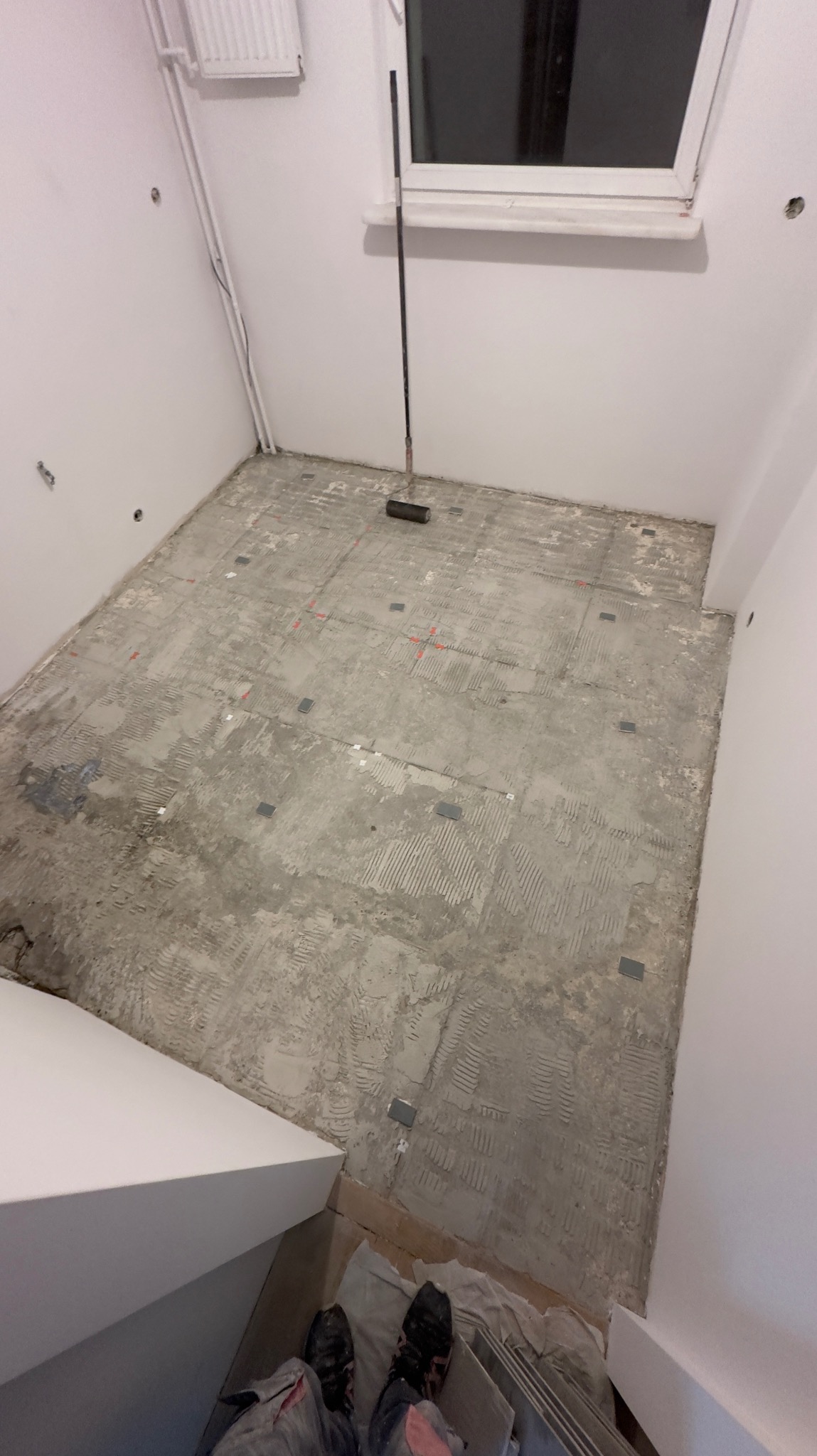 Przygotowanie podłoża pod płytki w łazience: widoczne ślady po kleju, poziomowanie, narzędzia i fragment osoby wykonującej prace.