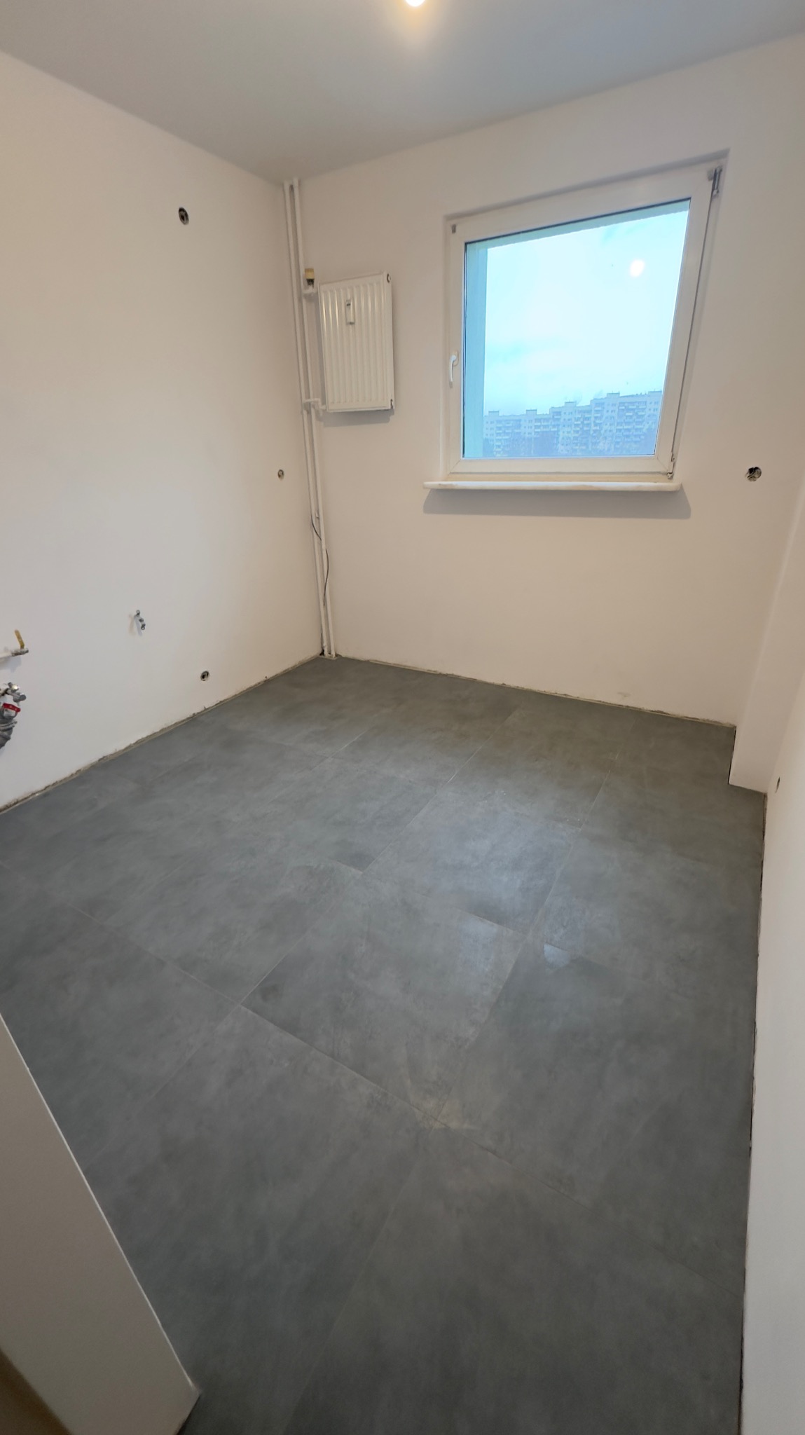Puste pomieszczenie po położeniu szarych płytek podłogowych, widoczny grzejnik na ścianie i okno z widokiem na budynki.