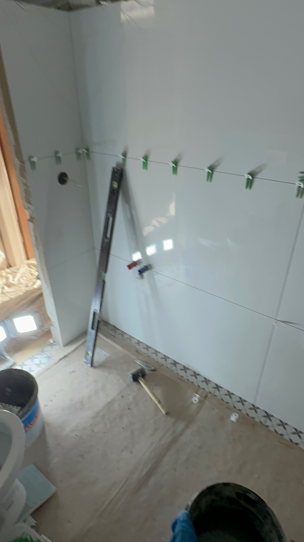 Świeżo ułożone, błyszczące, białe płytki na ścianie łazienki, wyrównane przy pomocy poziomicy i klipsów, widoczne narzędzia: młotek, wiadro z zaprawą, częściowo ułożona podłoga z dekoracyjnym pasem...