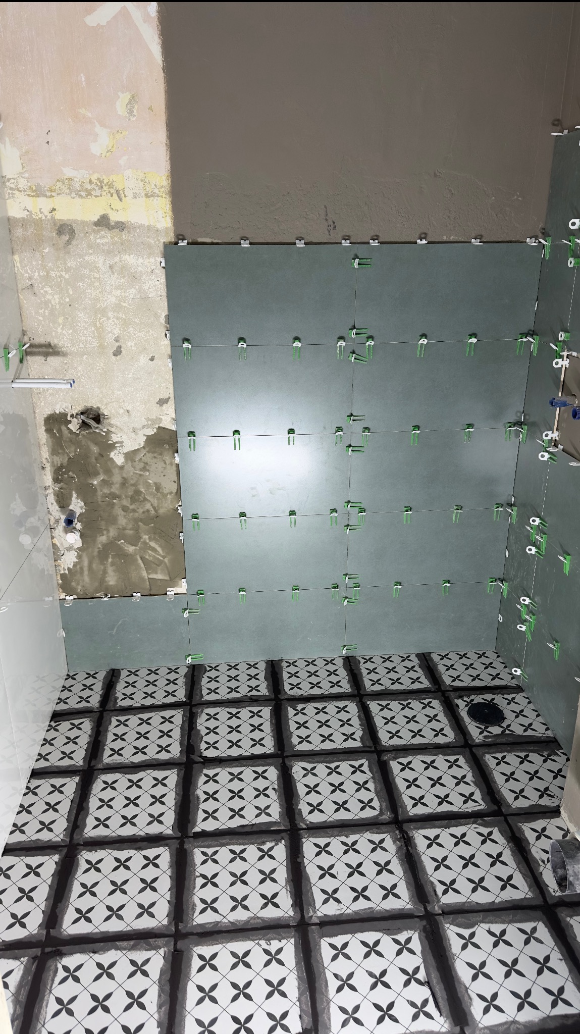 Świeżo ułożone, wzorzyste płytki podłogowe w łazience, z widocznym odpływem liniowym. Na ścianach szare płytki częściowo ułożone z użyciem klipsów poziomujących, oraz fragment ściany z widocznym...