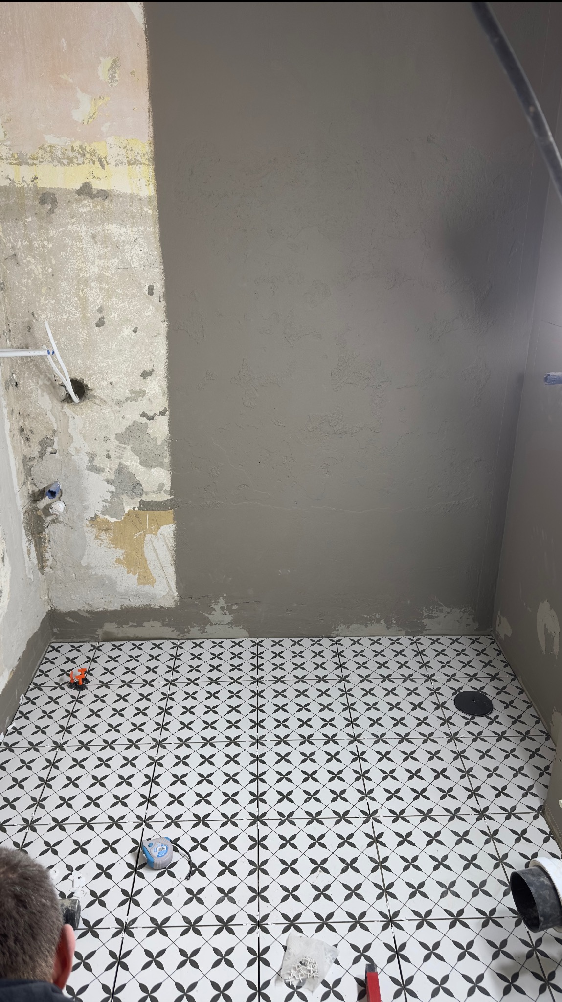 Łazienka w trakcie prac: ściana częściowo pokryta szarą gładzią, podłoga wyłożona biało-czarną mozaiką, widoczne narzędzia glazurnicze i fragment instalacji wodnej.