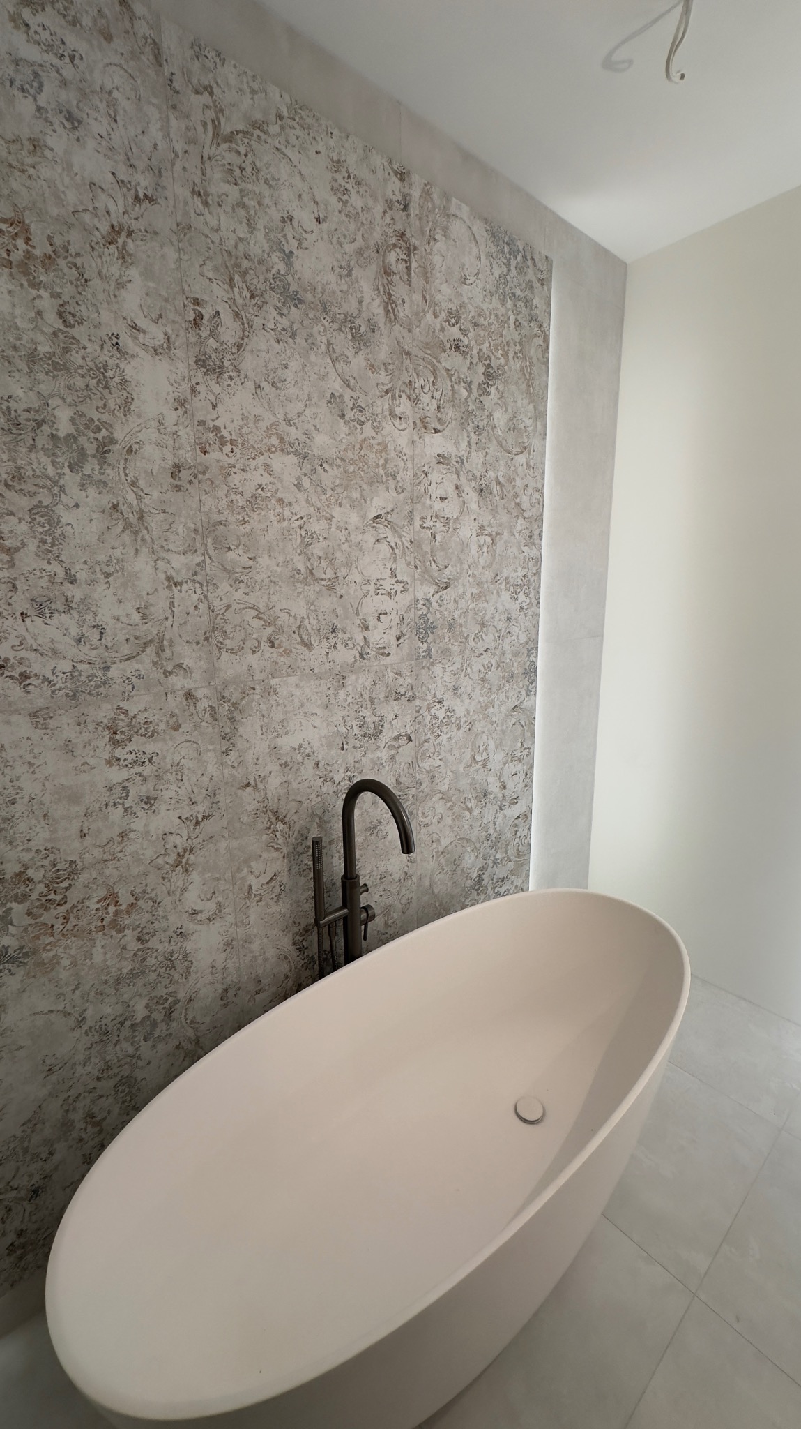 Wanna wolnostojąca o owalnym kształcie z czarną armaturą na tle ściany wyłożonej płytkami w stylu vintage, w jasnej łazience z szarą podłogą z płytek.