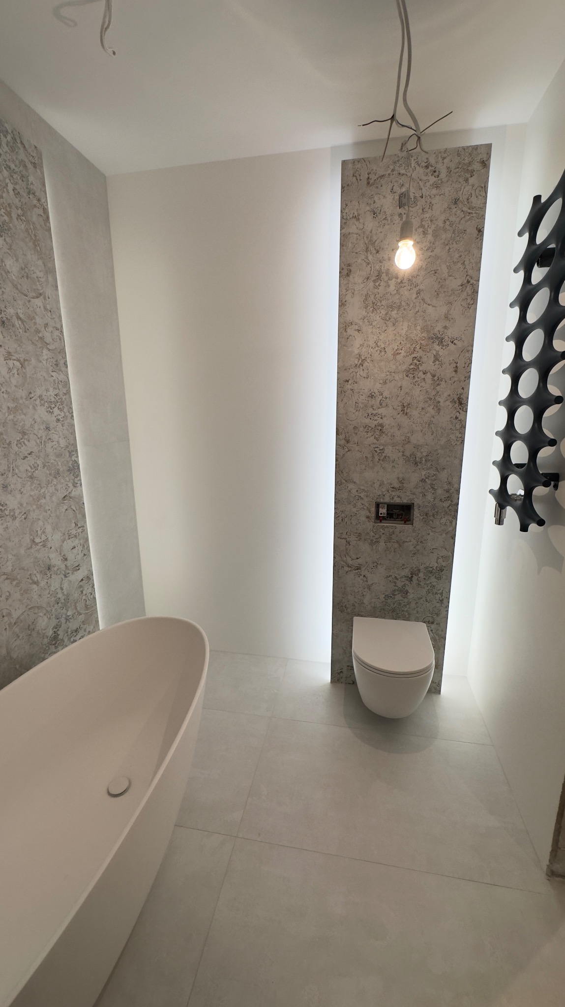Nowoczesna łazienka z wolnostojącą wanną, podwieszaną toaletą, dekoracyjnym grzejnikiem i panelami ściennymi o strukturze betonu, oświetlona wiszącą żarówką i ukrytym oświetleniem LED.