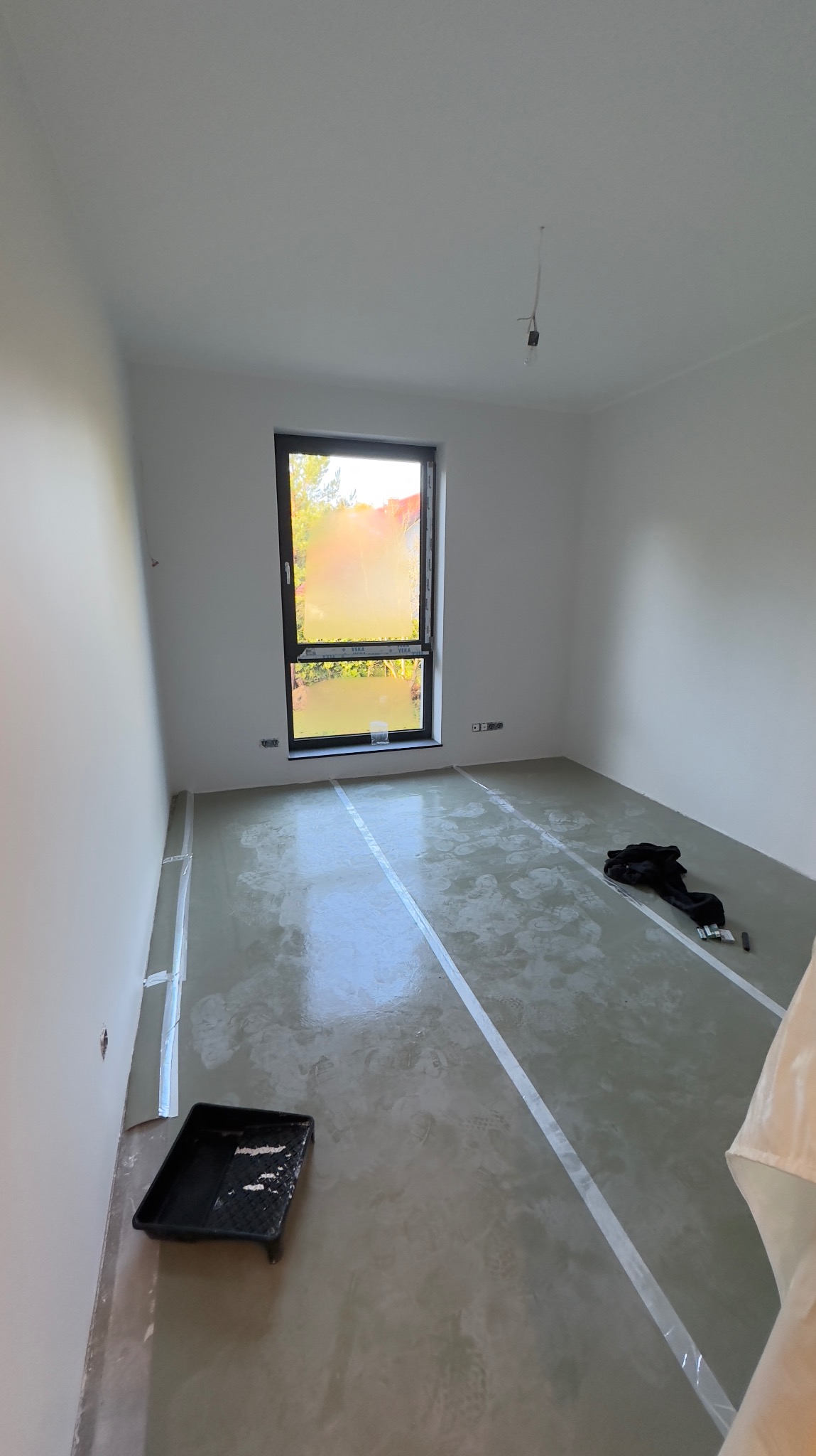 Przygotowane podłoże pod wylewkę samopoziomującą, zabezpieczone taśmą przy ścianach, widoczna kuweta malarska i ubrania robocze w pustym pomieszczeniu z oknem.