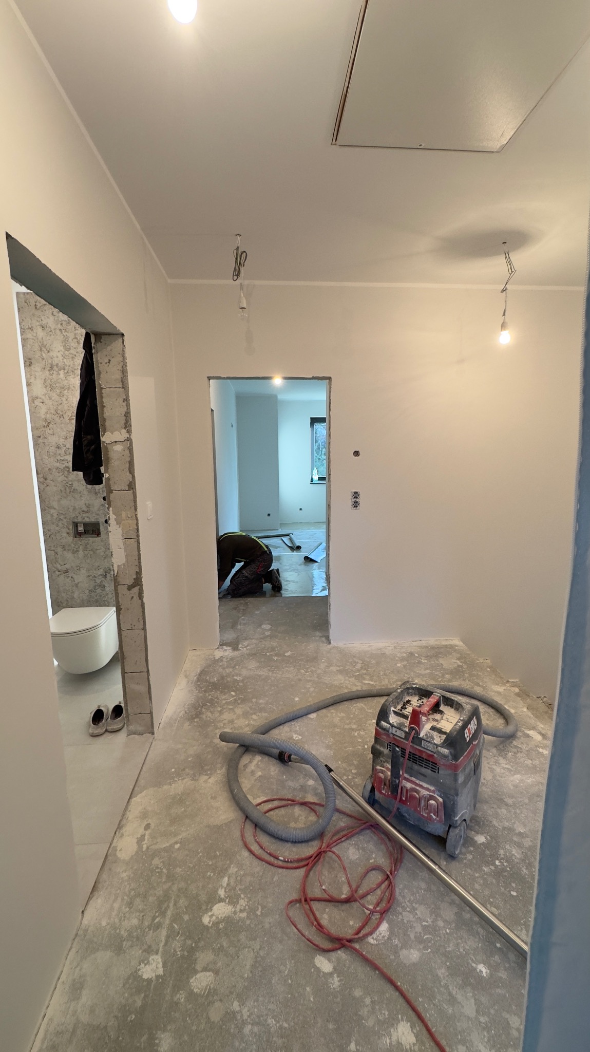Wnętrze w trakcie prac wykończeniowych: widoczny odkurzacz przemysłowy, surowy beton na podłodze, ściany po nałożeniu gładzi, oraz fragment łazienki z toaletą w tle. W głębi pomieszczenia, osoba...
