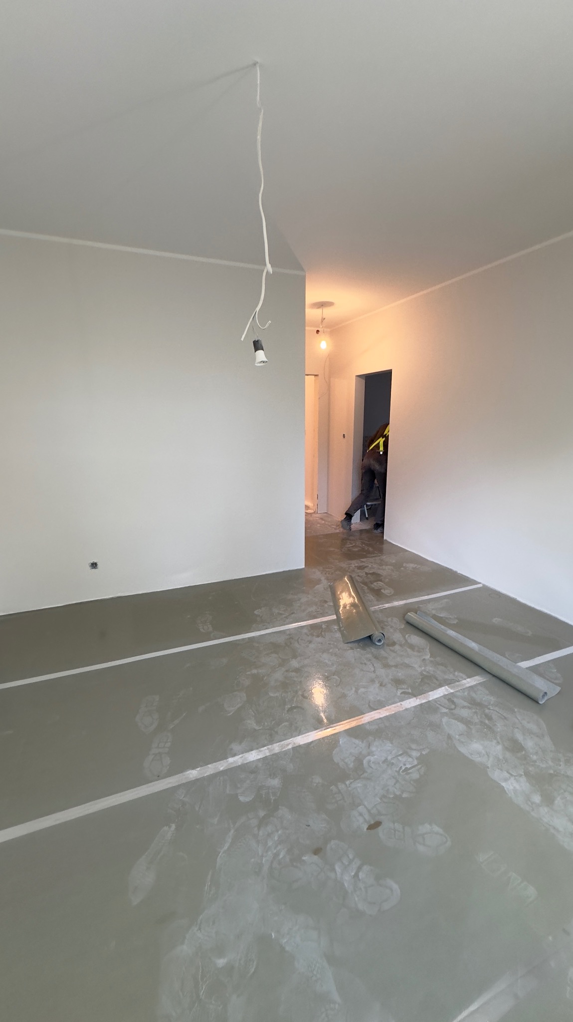 Przygotowanie do prac wykończeniowych w pustym pomieszczeniu, podłoga pokryta wylewką z widocznymi śladami butów, zabezpieczona folią malarską i taśmami, zwisający kabel z żarówką. W tle widoczna...