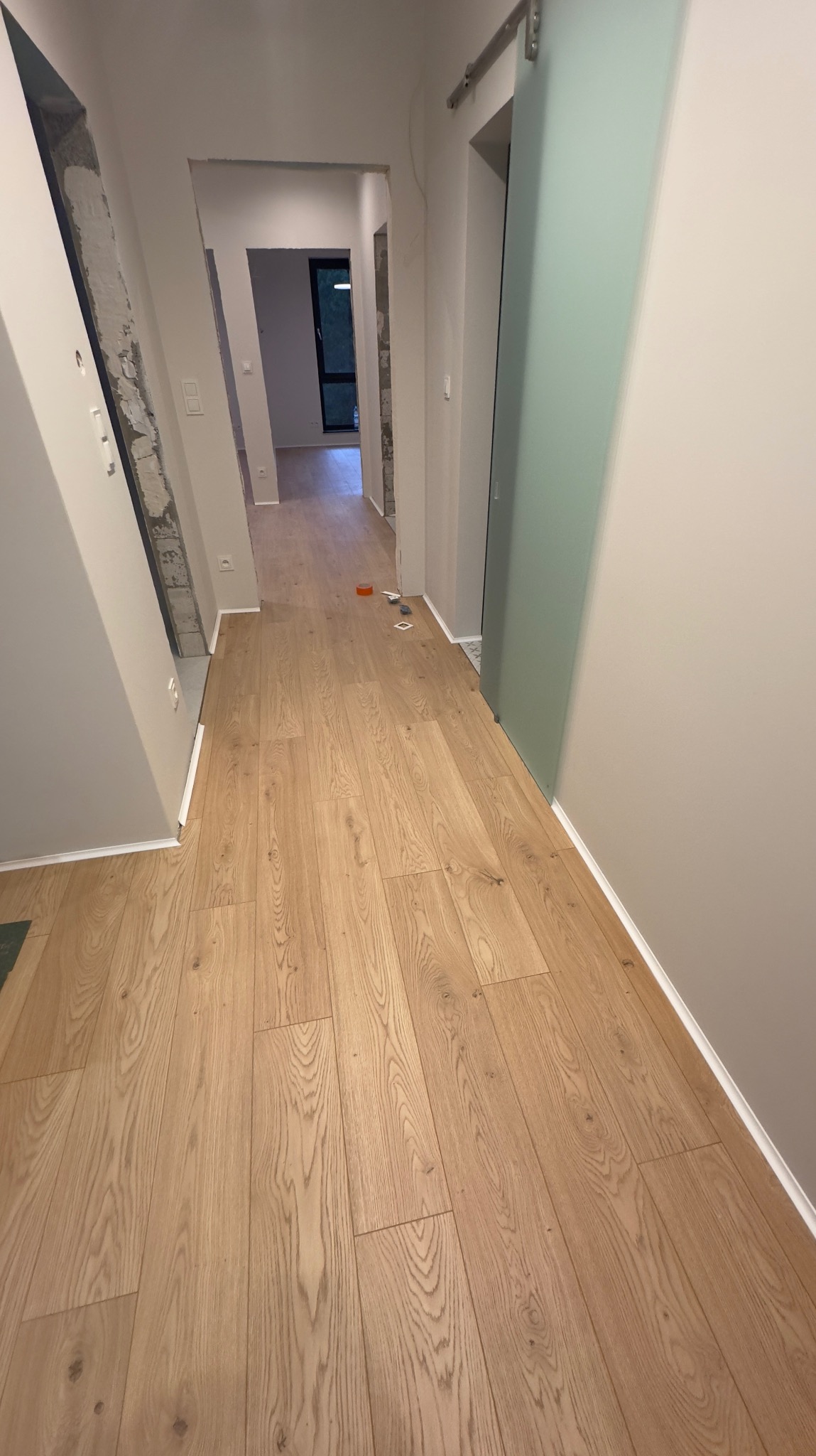 Nowo ułożona, jasna podłoga panelowa w korytarzu z widocznym fragmentem ściany w stanie surowym i zamontowanymi przesuwnymi drzwiami.