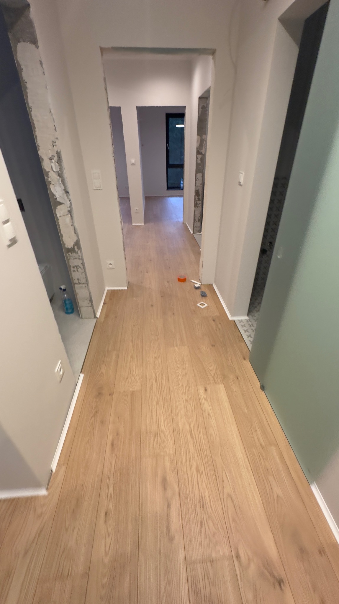 Korytarz w trakcie prac wykończeniowych, widoczne świeżo ułożone panele podłogowe, surowe ściany z śladami gładzi oraz narzędzia i akcesoria do montażu listew przypodłogowych.