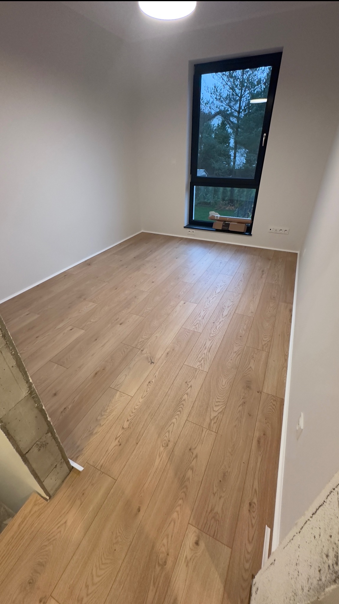 Jasne panele podłogowe ułożone w nowym pomieszczeniu z oknem, widoczne kartony pod oknem, surowy betonowy element konstrukcyjny w lewym dolnym rogu.