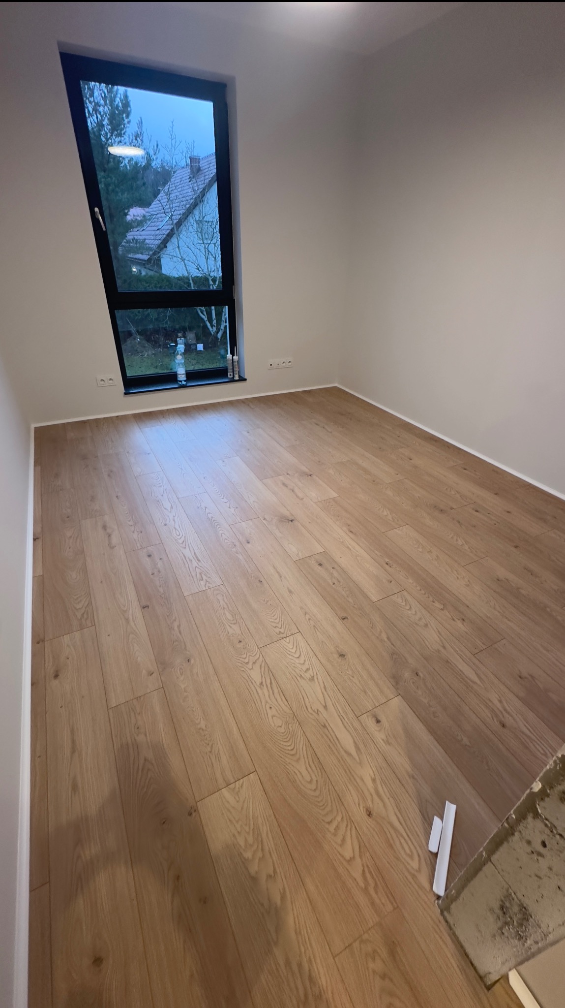 Jasne panele podłogowe ułożone w pokoju z oknem w kształcie trapezu, widok na dom z ogrodem, białe ściany, listwy przypodłogowe, butelka wody i silikon przy oknie.