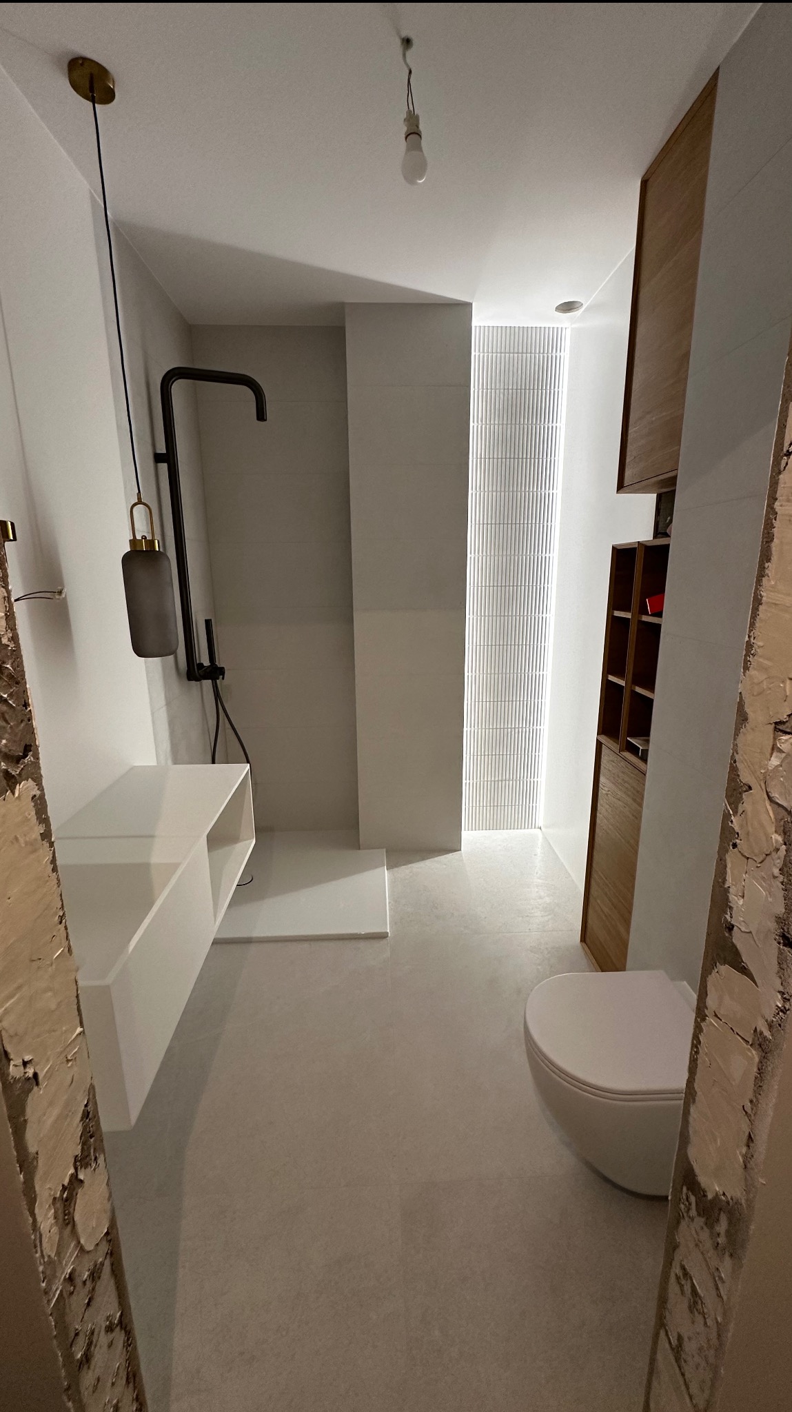 Nowoczesna łazienka w trakcie wykańczania, widoczna kabina prysznicowa typu walk-in, podwieszana umywalka z szafką, toaleta oraz pionowy panel dekoracyjny z podświetleniem, surowy stan wykończenia...