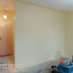 RaMo-BUD Rafał Budziszewski - Pokój z żółtymi ścianami w trakcie odświeżania, widoczne narzędzia malarskie i zabezpieczone drzwi folią malarską.