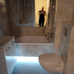 RaMo-BUD Rafał Budziszewski - Nowoczesna łazienka z szarymi marmurkowymi płytkami, podświetlaną wanną i wiszącą toaletą; odbicie fotografa w lustrze.