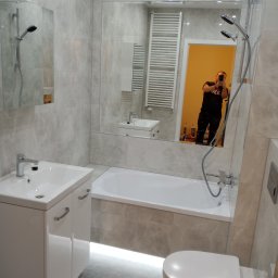 RaMo-BUD Rafał Budziszewski - Jasna łazienka z wanną, umywalką z szafką i toaletą, wykończona płytkami imitującymi marmur. W lustrze odbija się osoba robiąca zdjęcie.