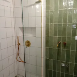 Nowoczesna kabina prysznicowa z białymi i zielonymi płytkami, złotą armaturą i odpływem liniowym w podłodze.