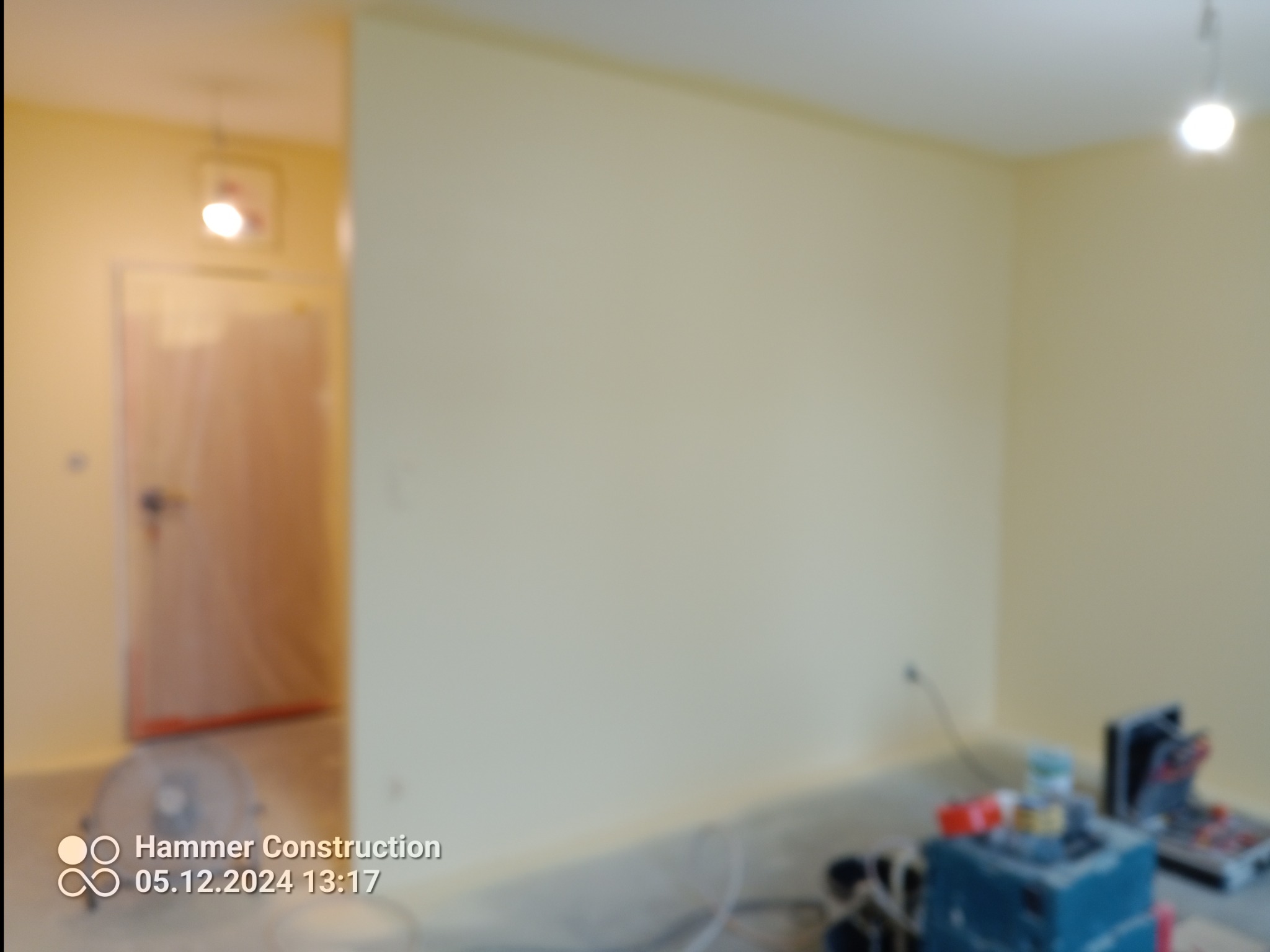 Pokój z żółtymi ścianami w trakcie odświeżania, widoczne narzędzia malarskie i zabezpieczone drzwi folią malarską.