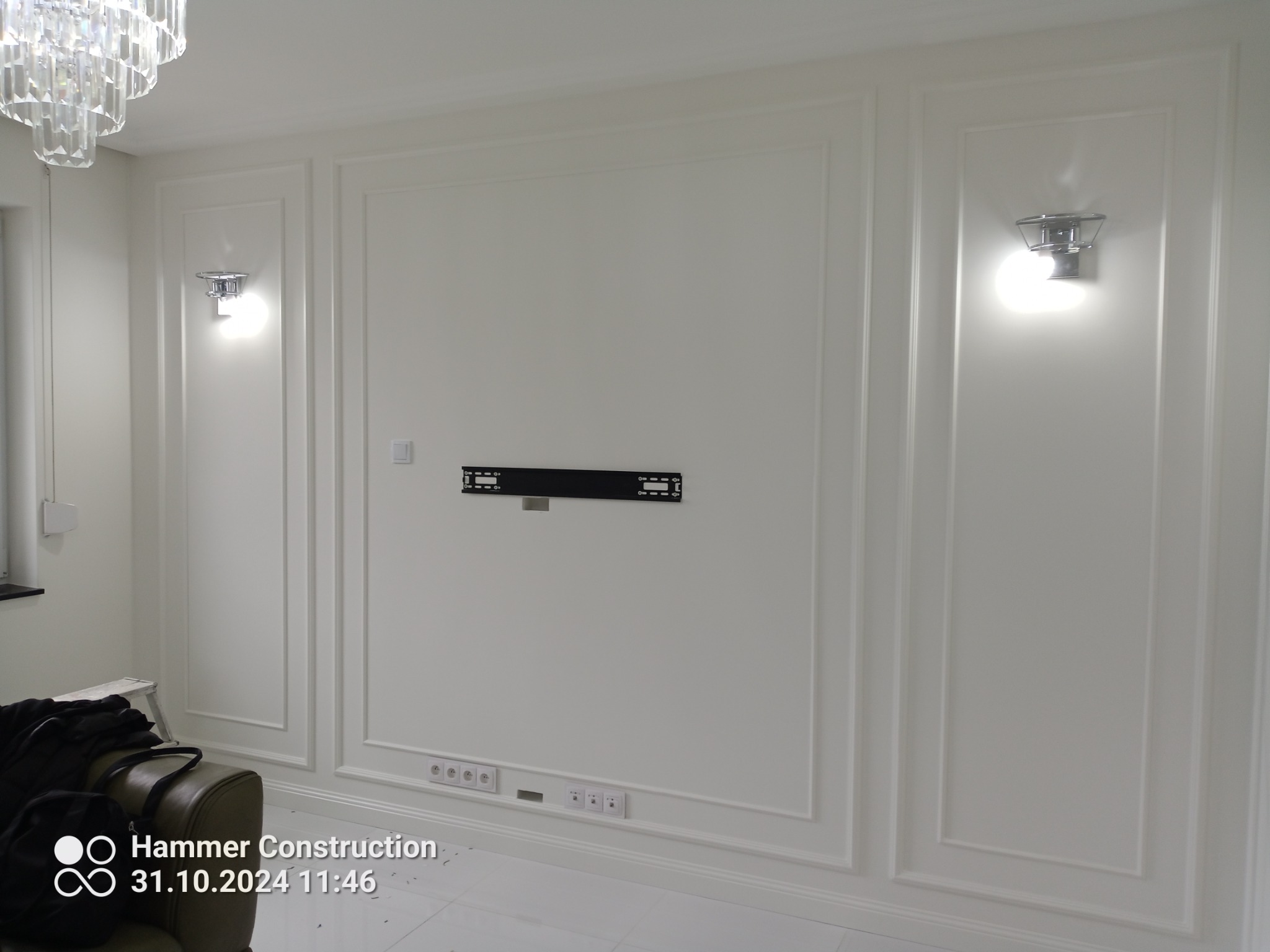 Eleganckie, białe panele ścienne z klasycznymi listwami dekoracyjnymi, oświetlone kinkietami i przygotowane do montażu telewizora. Widoczne gniazdka elektryczne i fragment kryształowego żyrandola.