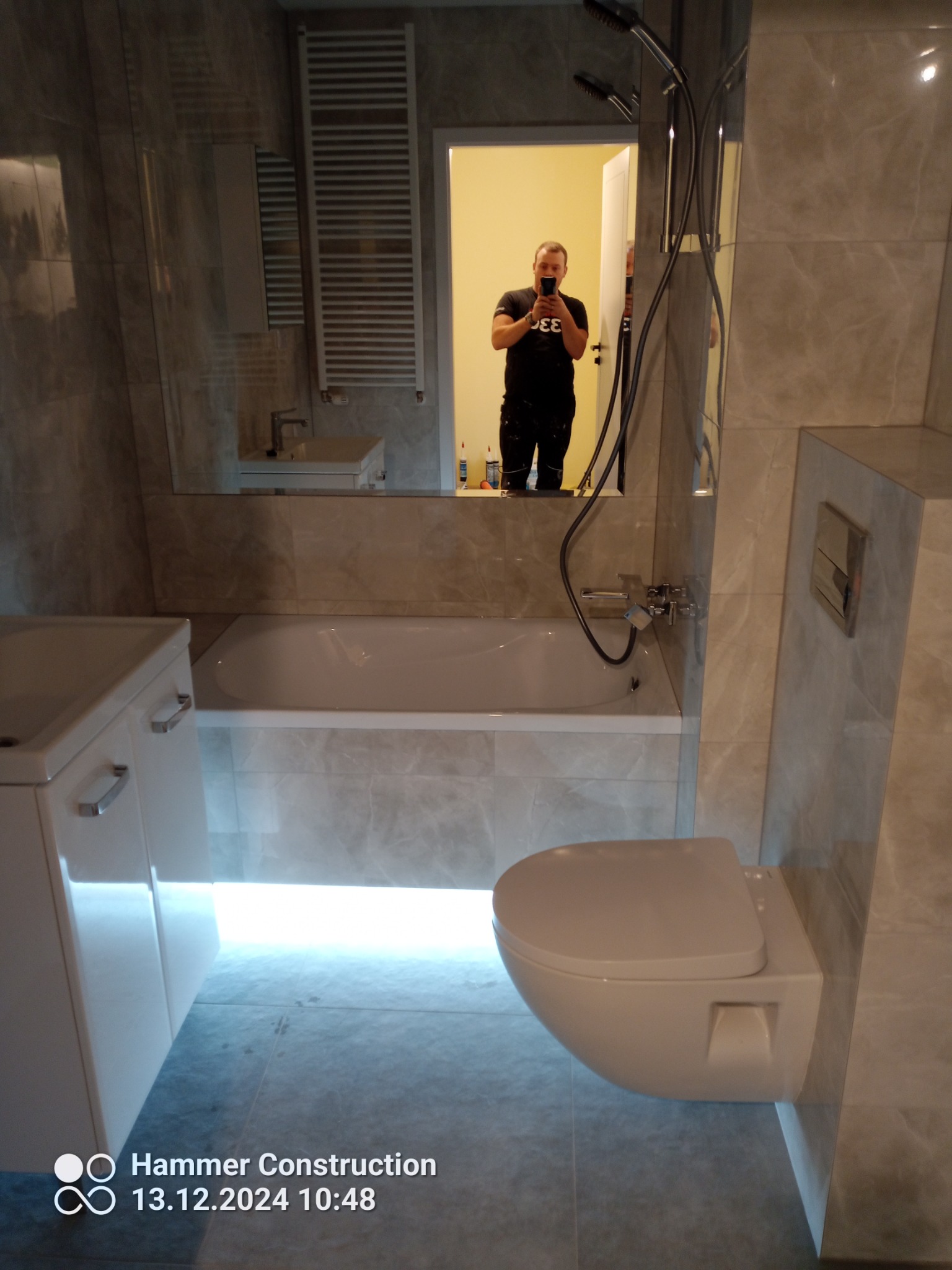 Nowoczesna łazienka z szarymi marmurkowymi płytkami, podświetlaną wanną i wiszącą toaletą; odbicie fotografa w lustrze.