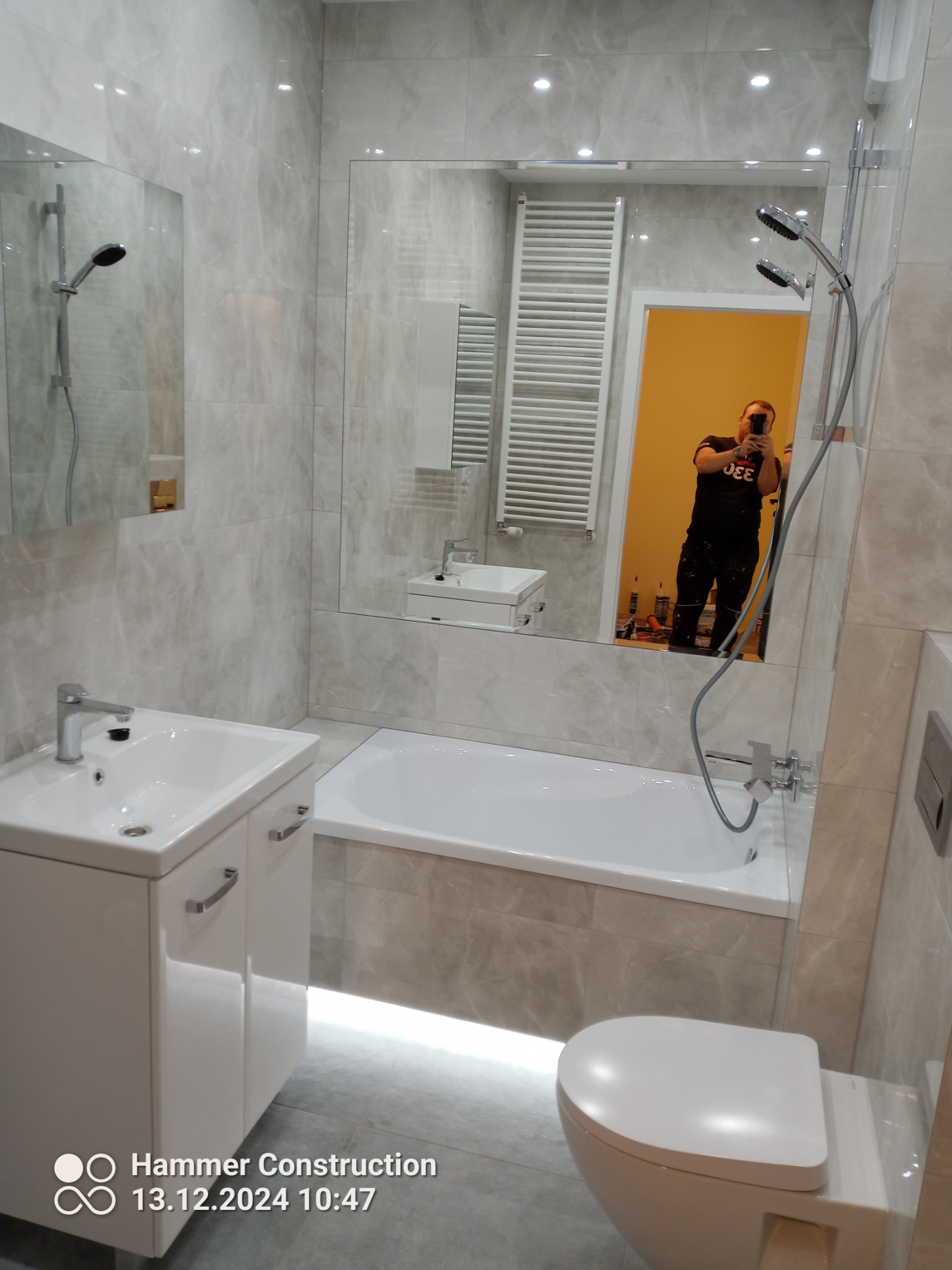 Jasna łazienka z wanną, umywalką z szafką i toaletą, wykończona płytkami imitującymi marmur. W lustrze odbija się osoba robiąca zdjęcie.