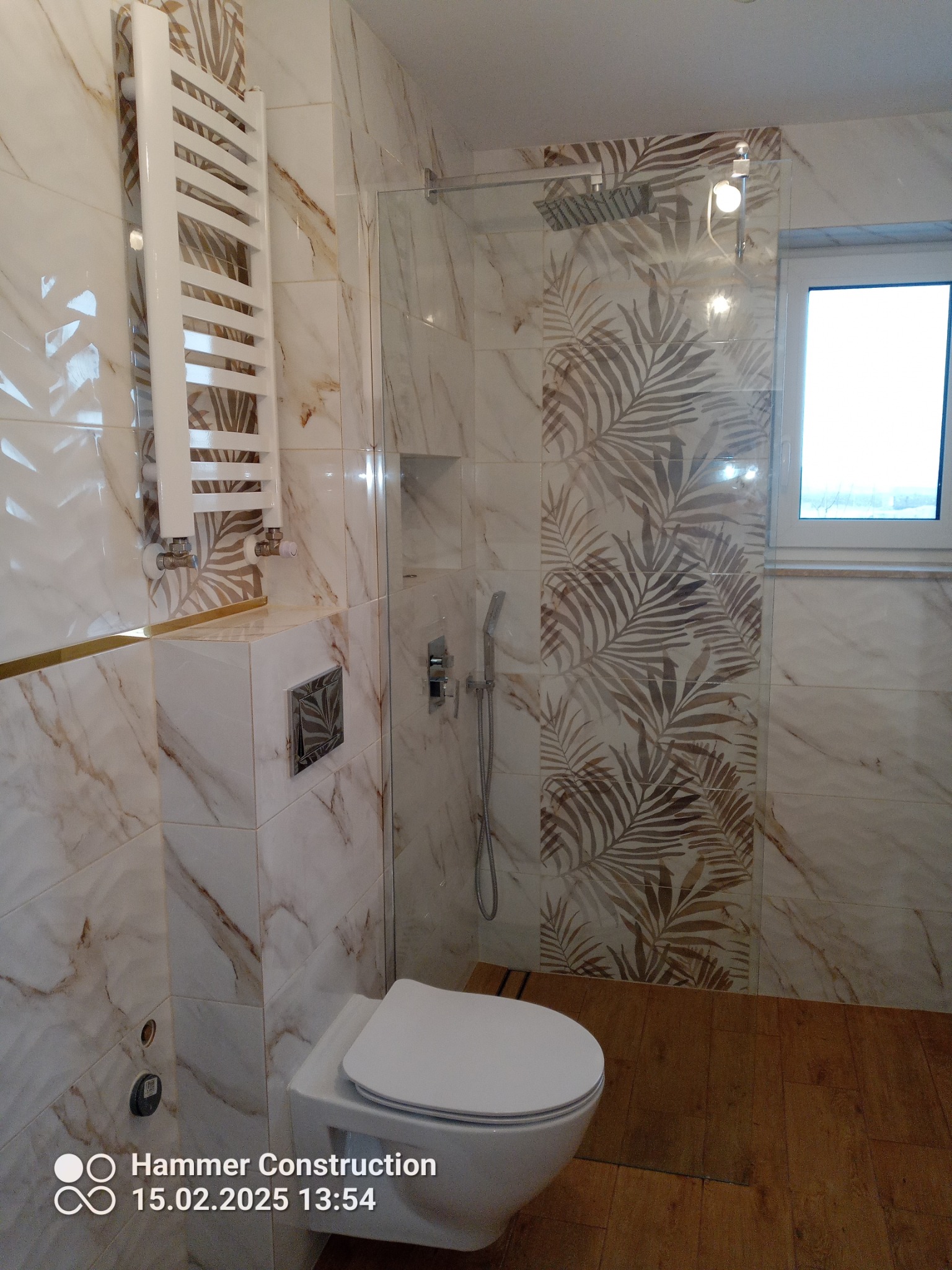 Nowoczesna łazienka z marmurowymi płytkami w odcieniach beżu i brązu z motywem liści, kabina prysznicowa ze szklaną ścianką, biały grzejnik drabinkowy i podłoga imitująca drewno.