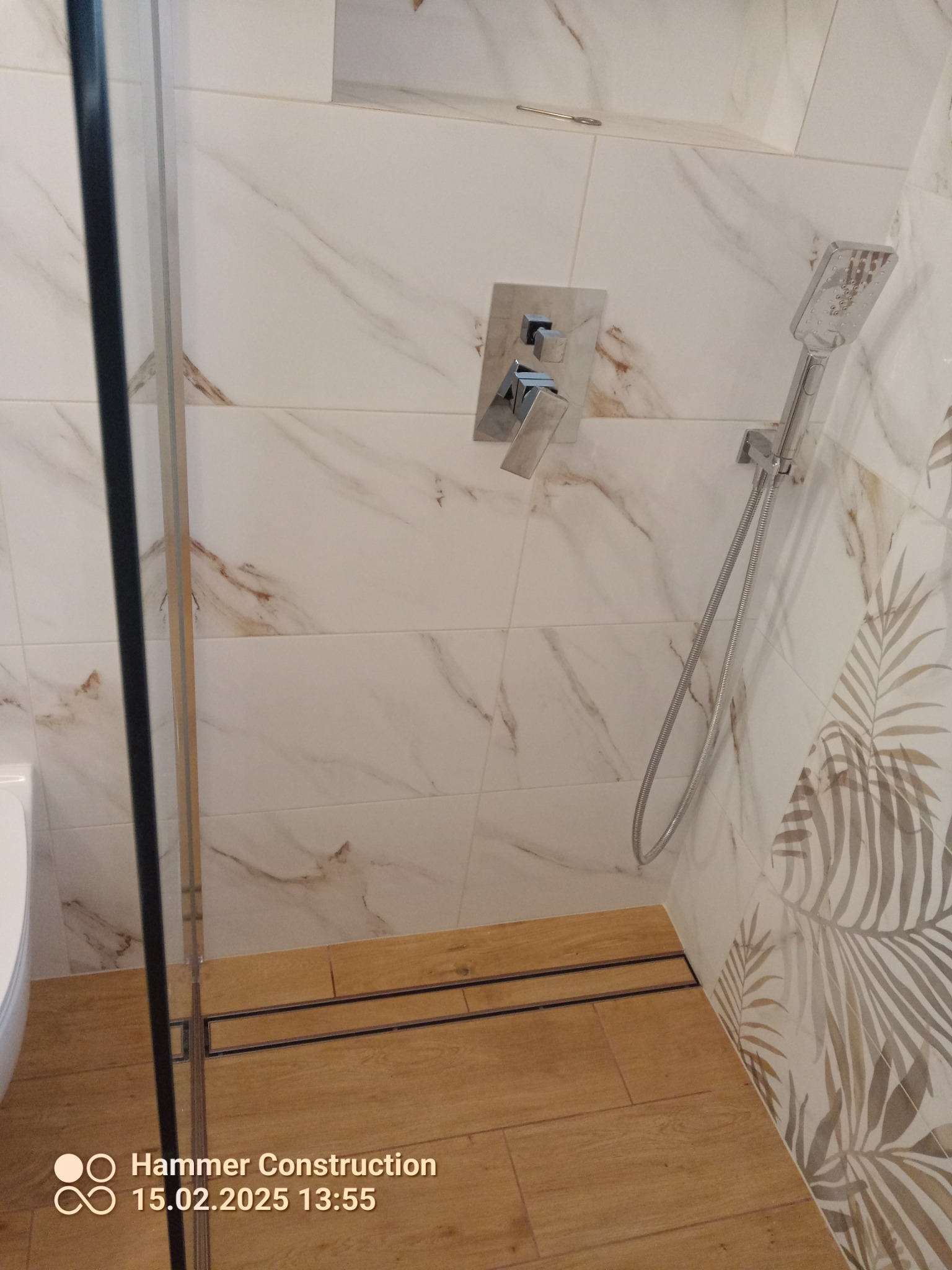 Nowoczesna kabina prysznicowa z odpływem liniowym, płytki imitujące marmur z delikatnym wzorem liści, chromowana bateria podtynkowa i słuchawka prysznicowa.