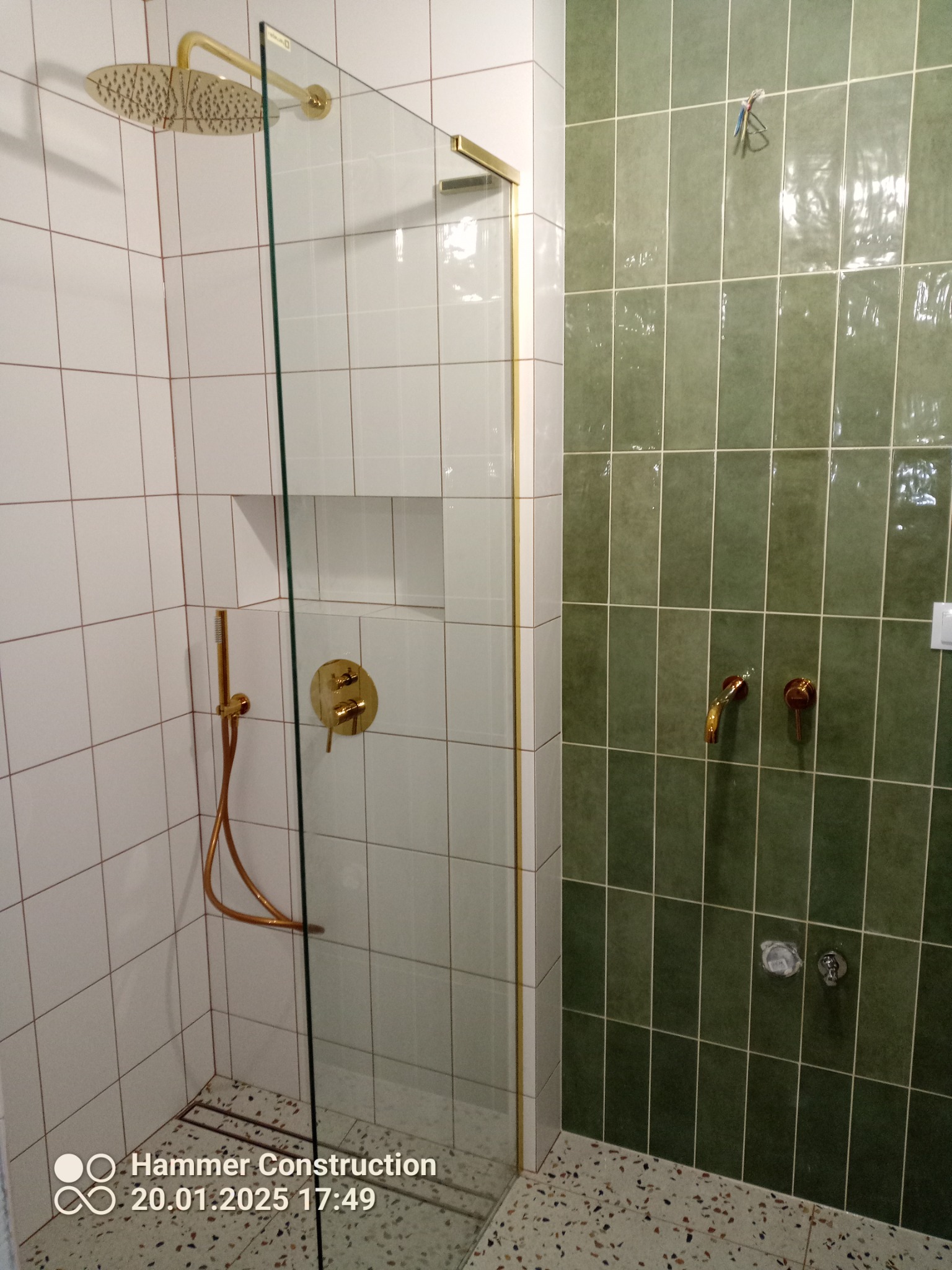 Nowoczesna kabina prysznicowa z białymi i zielonymi płytkami, złotą armaturą i odpływem liniowym w podłodze.