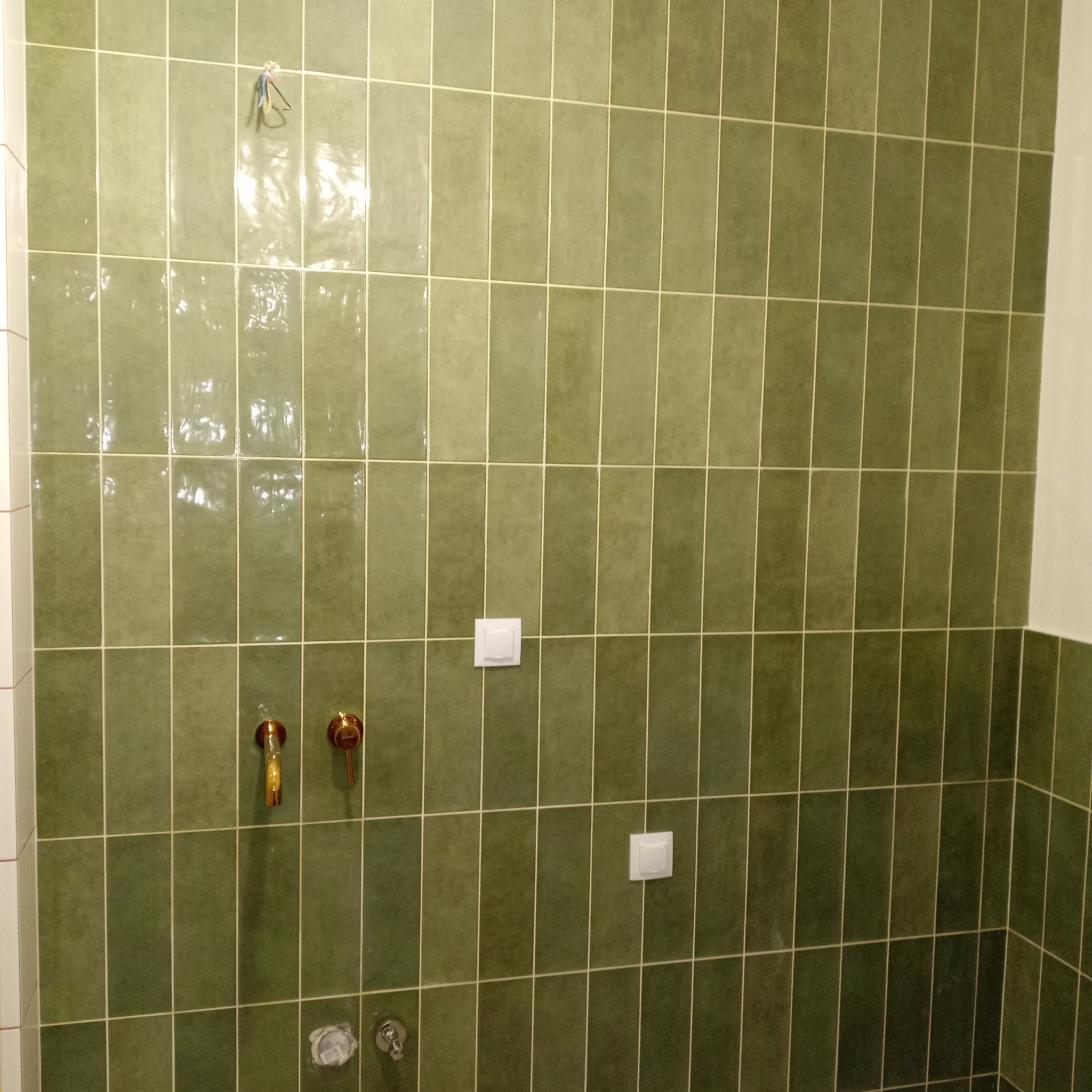 Ściana łazienki wyłożona pionowymi, oliwkowymi płytkami z jasną fugą, widoczne złote baterie, dwa białe włączniki światła i wystające kable.