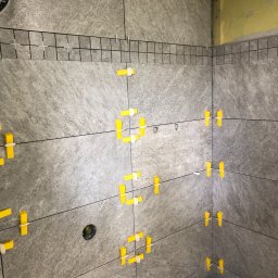 Piotr Kułagowski - Ściana w trakcie układania szarych płytek imitujących kamień, z widocznymi żółtymi krzyżykami dystansowymi i listwą wyrównującą na górze, przy niezabezpieczonym rogu ściany.