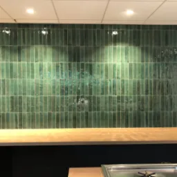 Ściana wyłożona pionowymi, błyszczącymi, zielonymi płytkami ceramicznymi z efektem postarzenia, oświetlona punktowo, z widocznym blatem roboczym poniżej i fragmentem wyposażenia kuchni w dolnej...