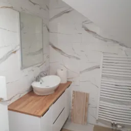 Wykończona łazienka na poddaszu: białe płytki imitujące marmur, drewnopodobny blat z owalną umywalką i grzejnik drabinkowy. Ukośne ściany.