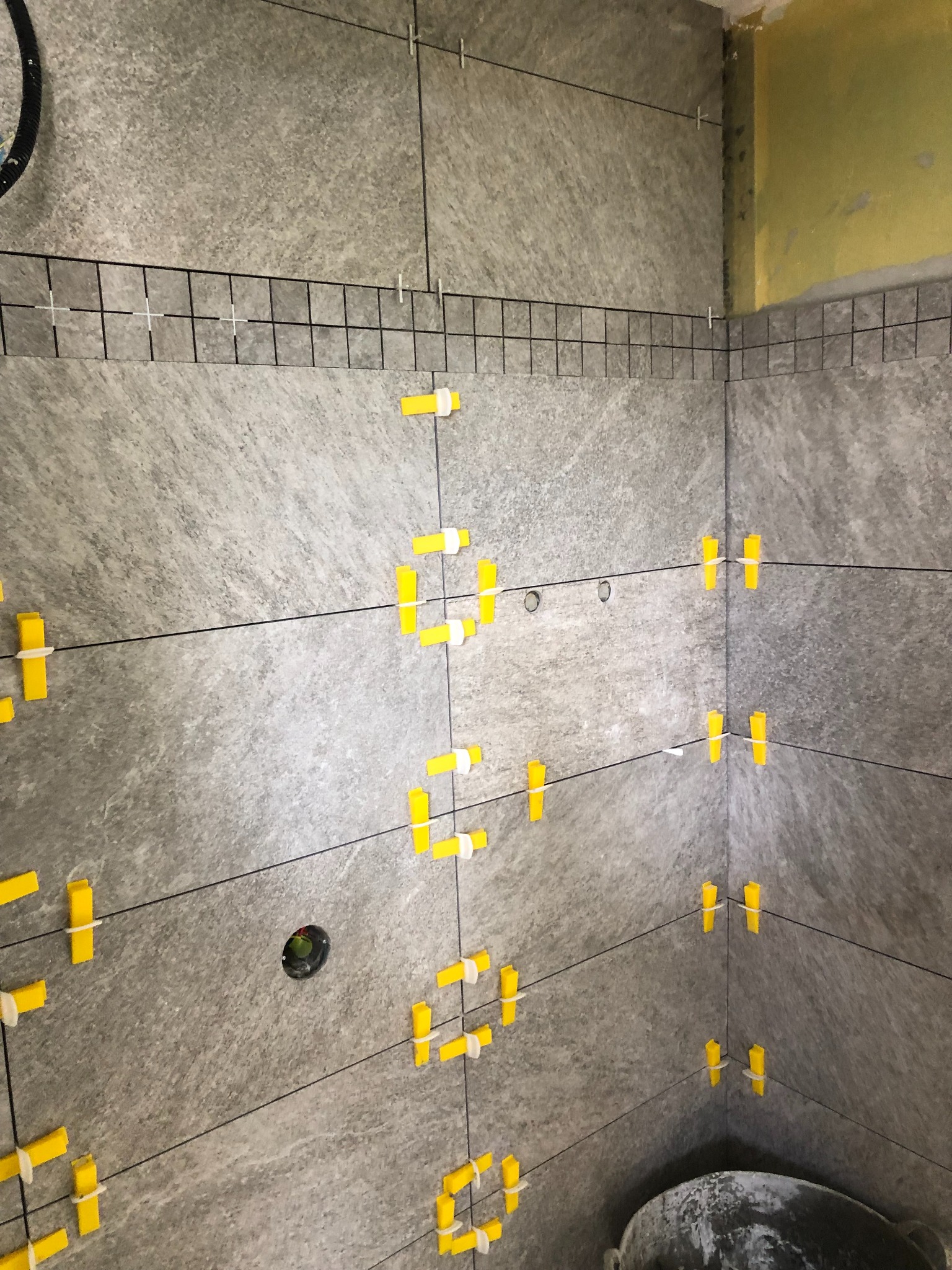 Ściana w trakcie układania szarych płytek imitujących kamień, z widocznymi żółtymi krzyżykami dystansowymi i listwą wyrównującą na górze, przy niezabezpieczonym rogu ściany.