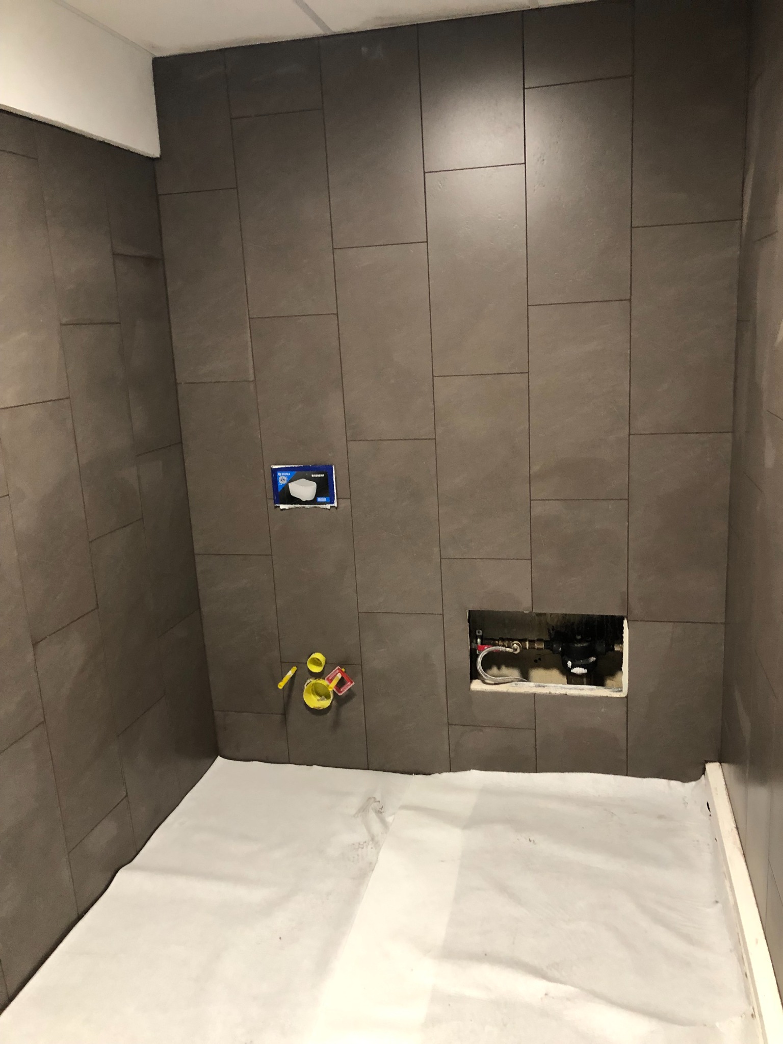 Ściany łazienki w trakcie układania brązowych płytek, widoczne instalacje wodne i kanalizacyjne, zabezpieczona podłoga, akcesoria glazurnicze.