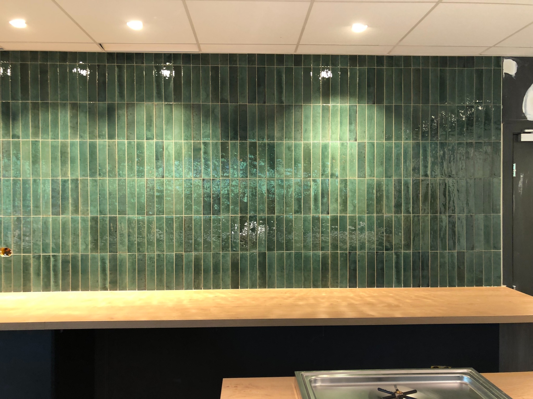 Ściana wyłożona pionowymi, błyszczącymi, zielonymi płytkami ceramicznymi z efektem postarzenia, oświetlona punktowo, z widocznym blatem roboczym poniżej i fragmentem wyposażenia kuchni w dolnej...