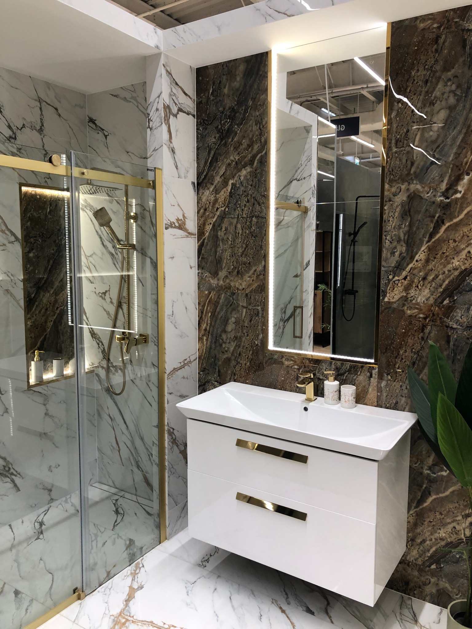 Nowoczesna łazienka z kabiną prysznicową ze złotymi detalami, białą umywalką z szafką i podświetlanym lustrem na tle marmurowych ścian w odcieniach bieli i brązu.